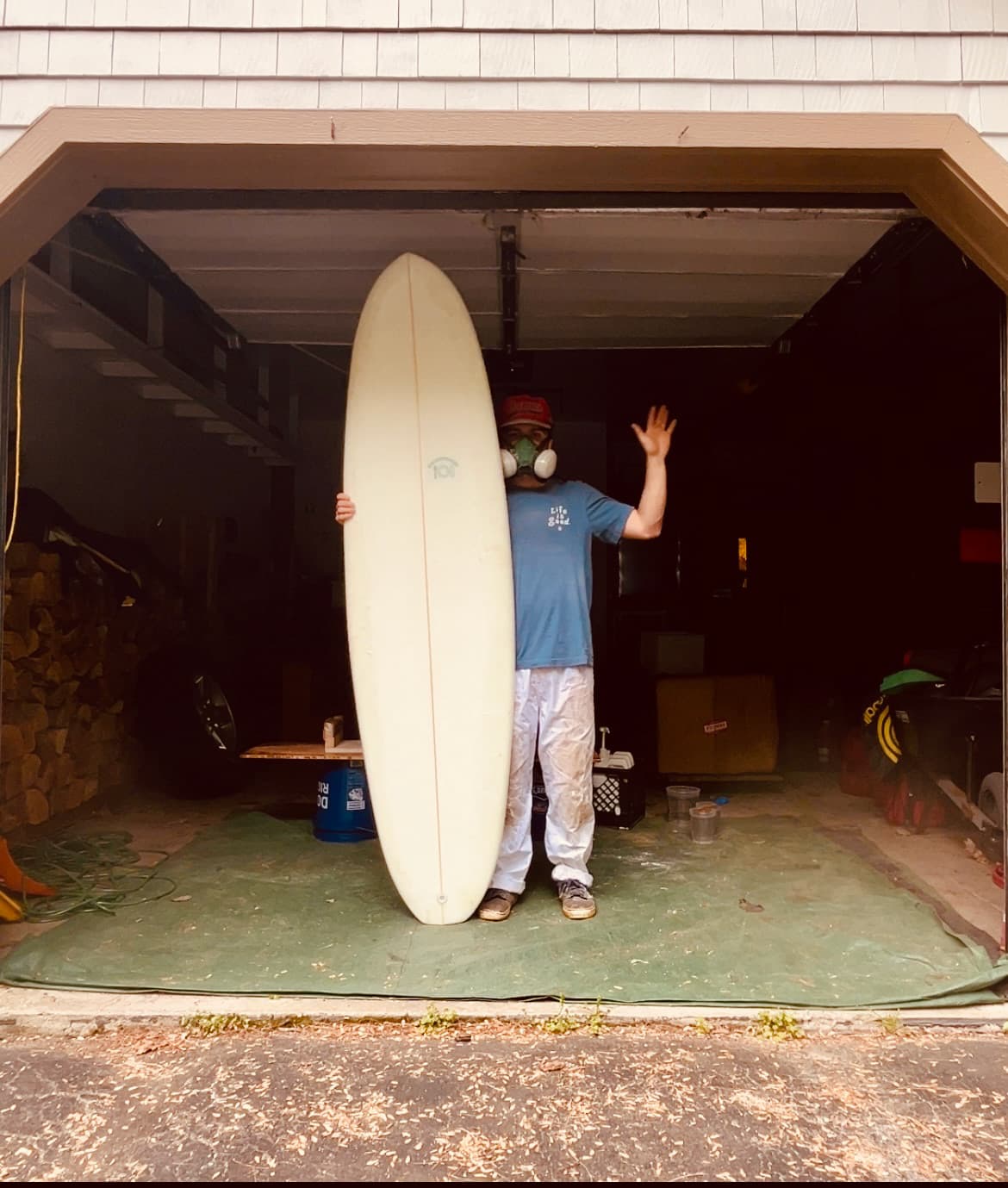 Kyle waving from a garage doorway beside a tall white Dish surfboard
