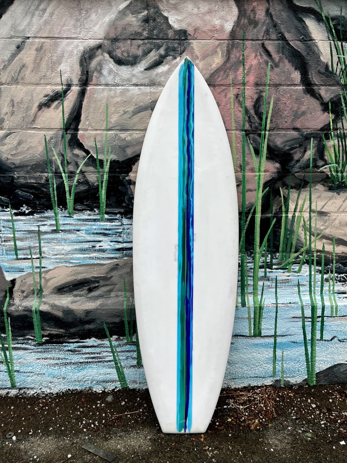 A white Dish surfboard with a blue stripe leaning against a painted mural